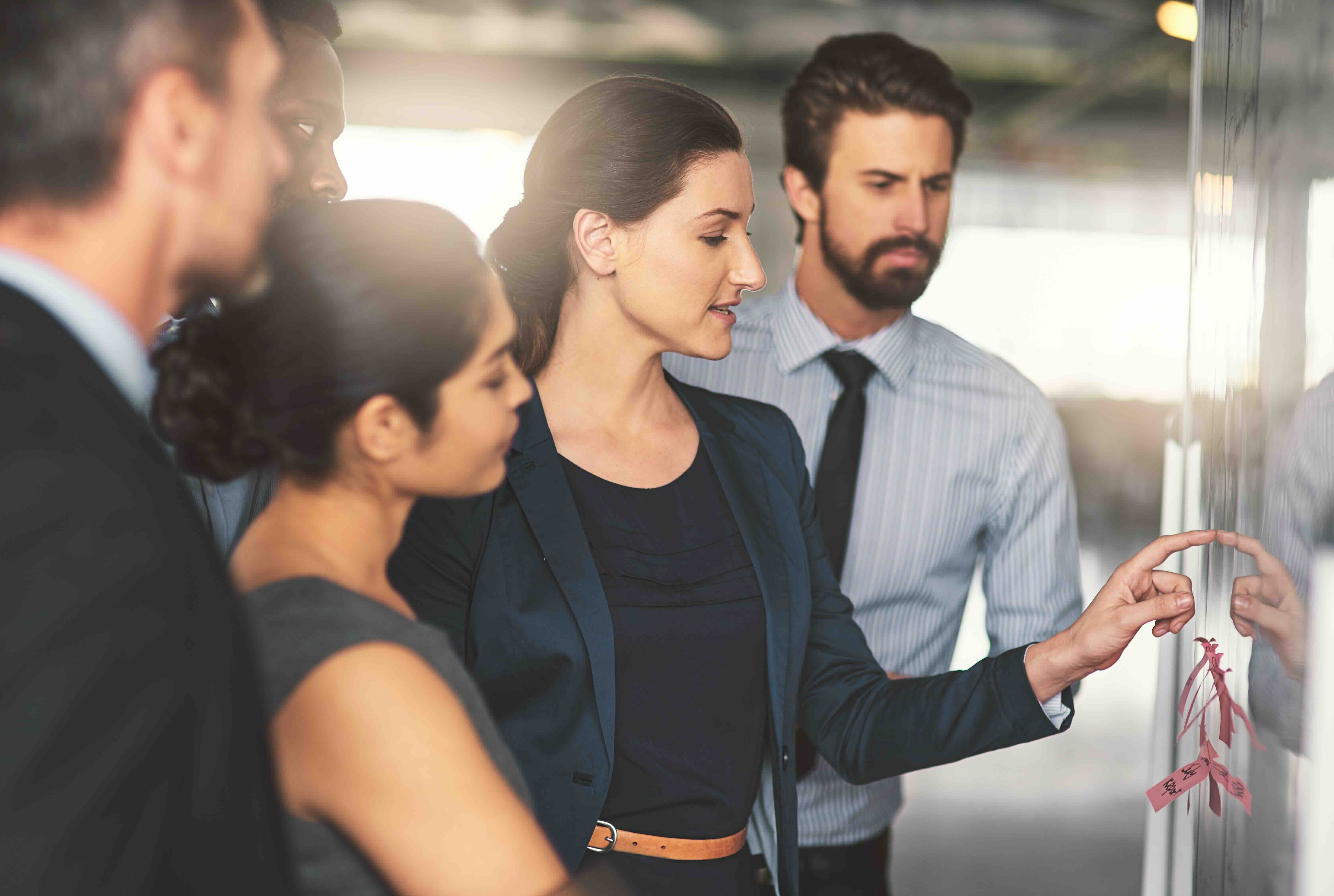 Business professionals gathered around woman pointing at organizational chart on glass board