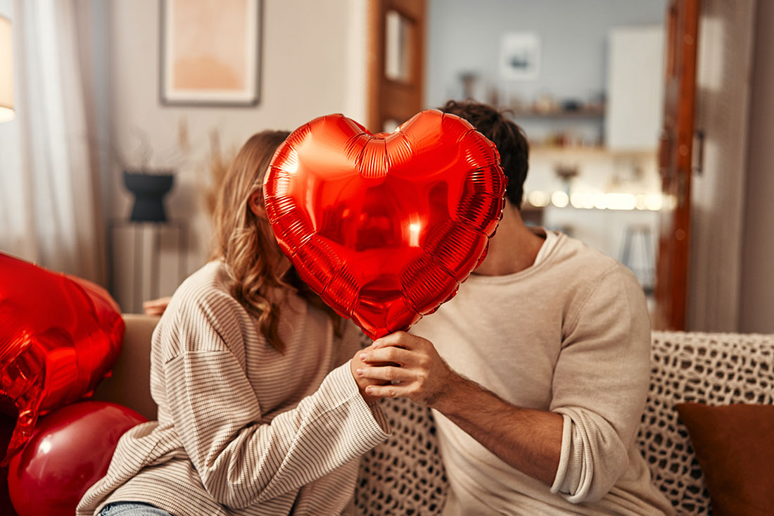 Young couple in love holding a heart-shaped balloon, covering themselves with it while kissing, sitting on the sofa in the living room at home.