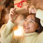 Asian woman in a white turtleneck smiling because she's holding gifts for people who work from home above her head