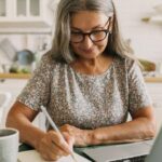 Photograph of a mature white woman with long grey hair and black thick rimmed glasses looking down and doing a personal swat analysis at her kitchen table.