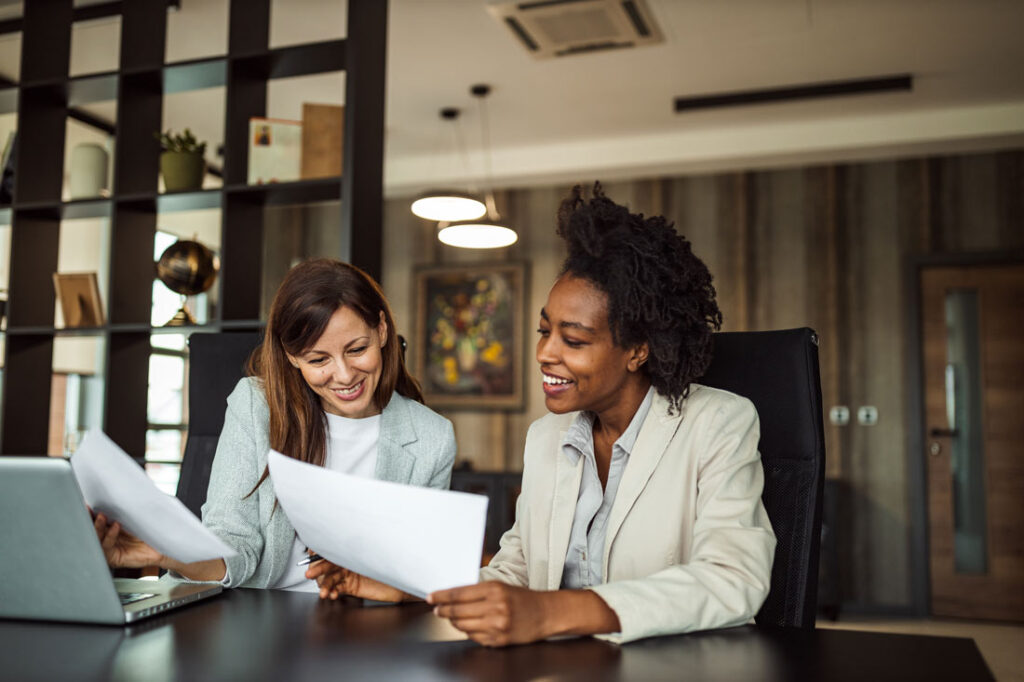 Two business women, on the left a white woman with long brown hair, on the right a Black woman with curly black hair, sitting at a conference table, working together and smiling showing the importance of emotional intelligence in the workplace.