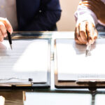 two hands holding pens, each hovering over paperwork to decide between an agreement vs contract