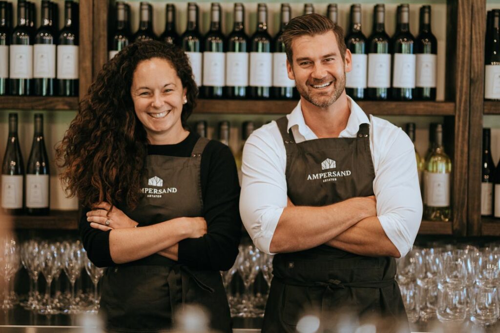 Photograph of the founders of Ampersand Estates, Melissa Bell and Corrie Scheepers, smiling and wearing aprons in front of a shelf of wine bottles
