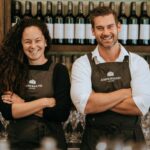 Photograph of the founders of Ampersand Estates, Melissa Bell and Corrie Scheepers, smiling and wearing aprons in front of a shelf of wine bottles