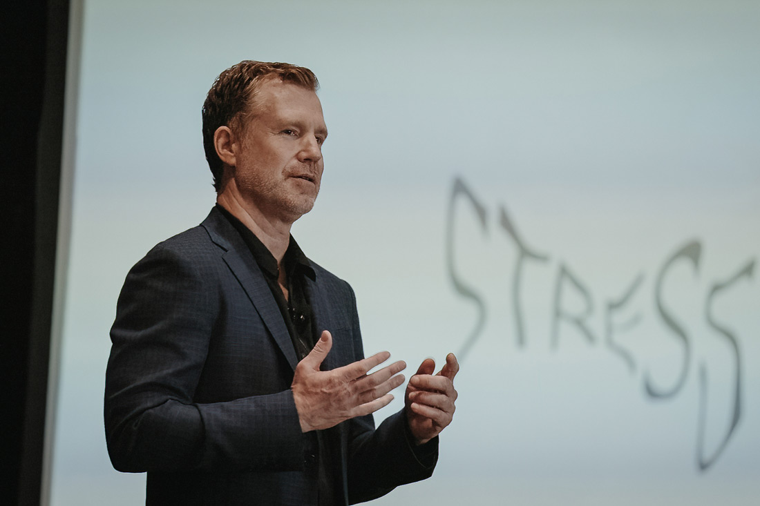 Photograph of a white man in a grey suit speaking with his hands in front of a white board that says "stress"