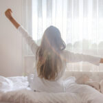 Photograph of a woman with long brown hair from behind sitting on her bed in her pajamas stretching her arms in front of a bright window to demonstrate she is a morning person