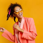 Photograph of an African American woman smiling and dancing in a salmon pink suit and yellow sunglasses to represent dopamine dressing