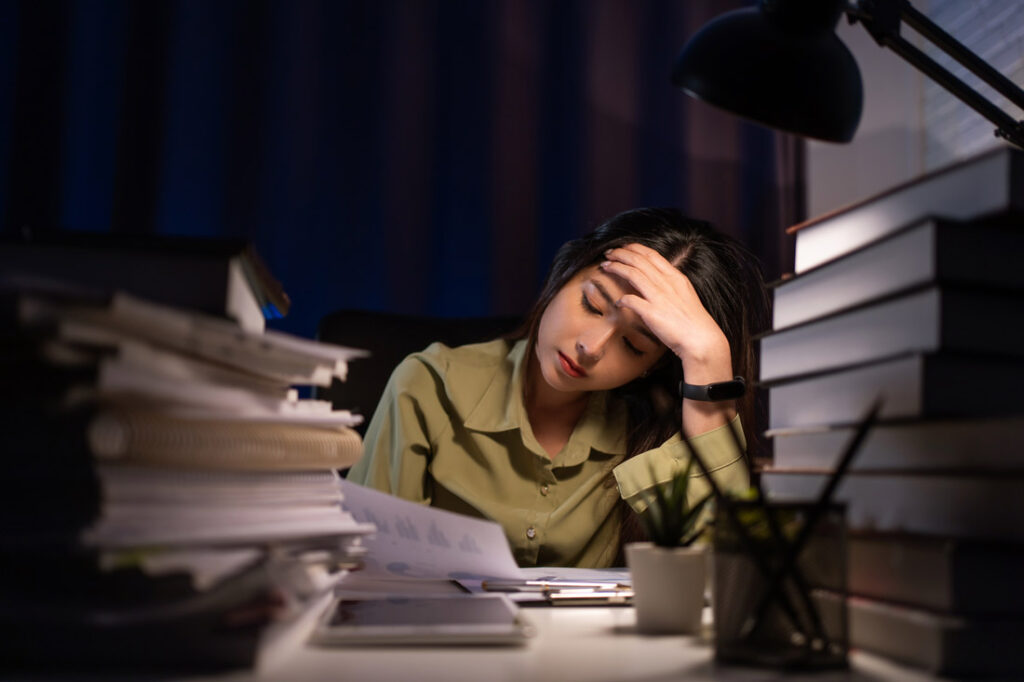 Young Asian woman leaning over desk surrounded by books late at night stuck in the performance paradox