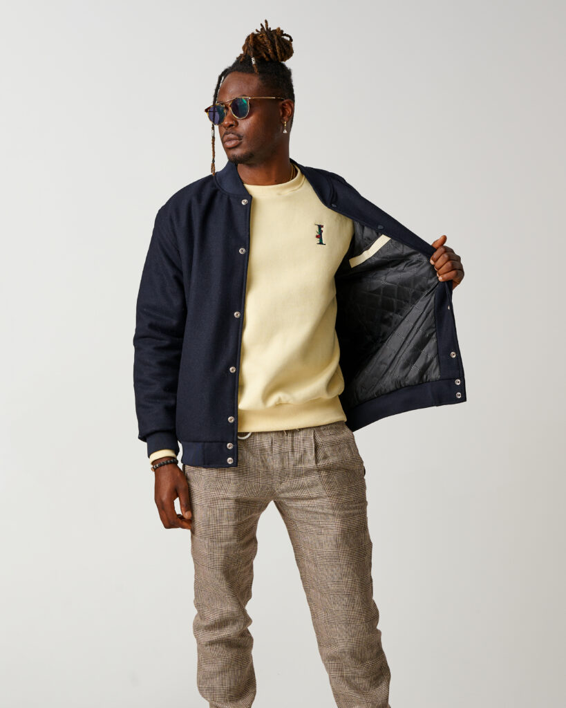 INHERENT model wearing sunglasses, a yellow sweatshirt and a navy blue letterman jacket