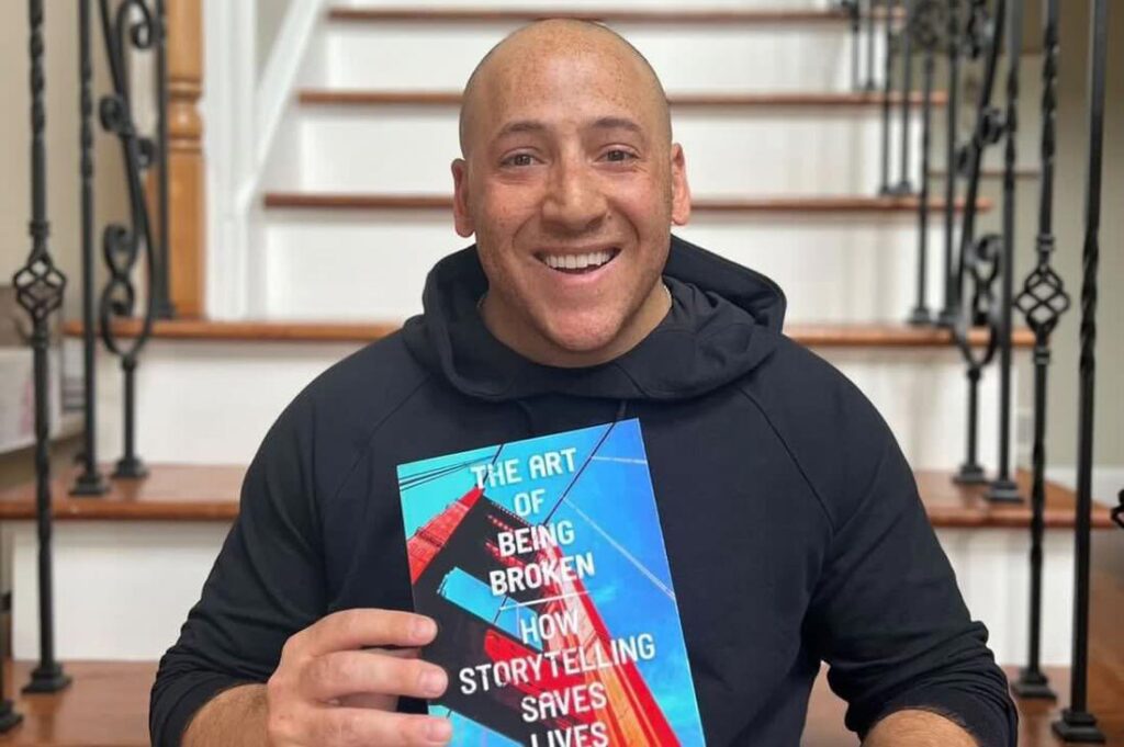 Kevin Hines sitting on a staircase with his new book "the art of being broken how storytelling saves lives"