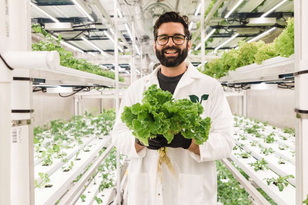 Dylan McCart, executive director of churchill northern studies centre, in a white lab coat holding fresh lettuce inside the Rocket Greens shipping container, surrounded by hydroponic leafy greens.