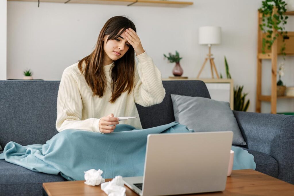 Sick businesswoman holding a thermometer in front of her laptop experiencing sick day guilt