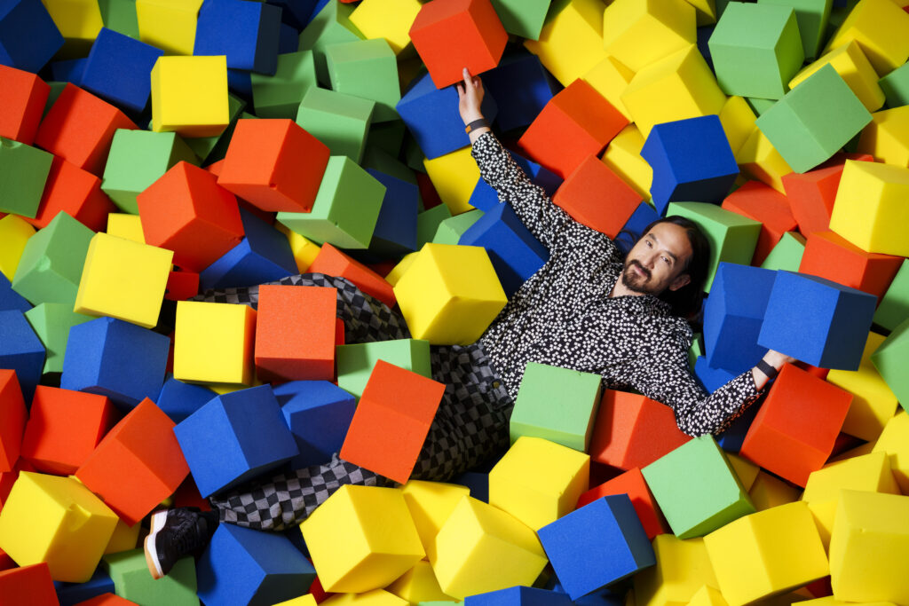 Steve Aoki laying in a colorful styrofoam ball pit. Photo by Eric Jamison Studio.