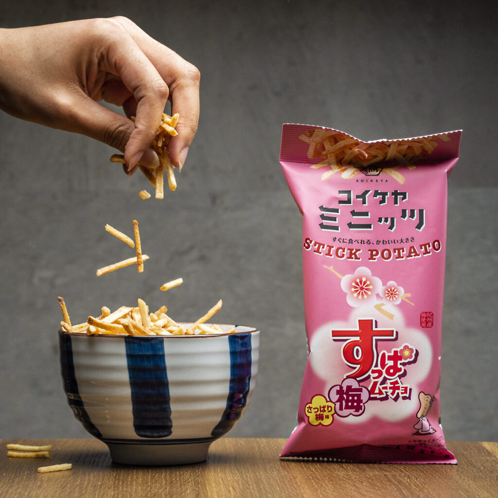 product image of hand dropping potato stick snacks into a ceramic bowl next to a bright pink bag of the potato sticks