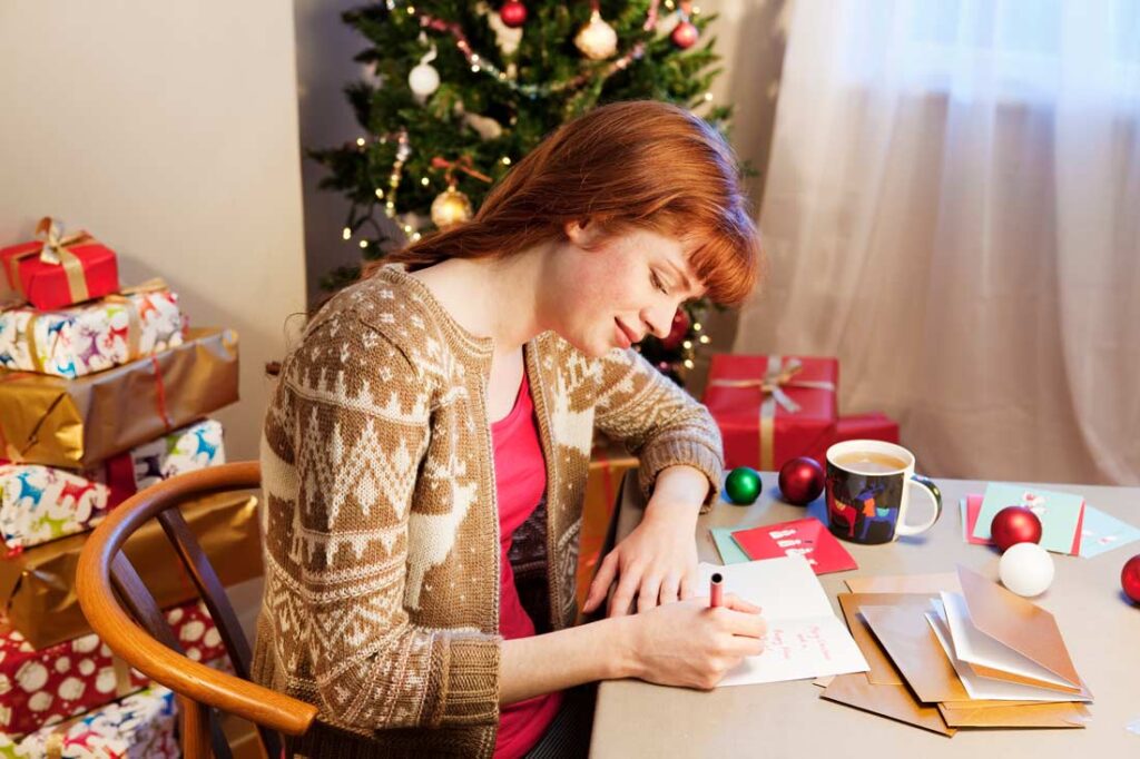 woman sitting at desk writing christmas cards and wondering how to politely stop sending christmas cards