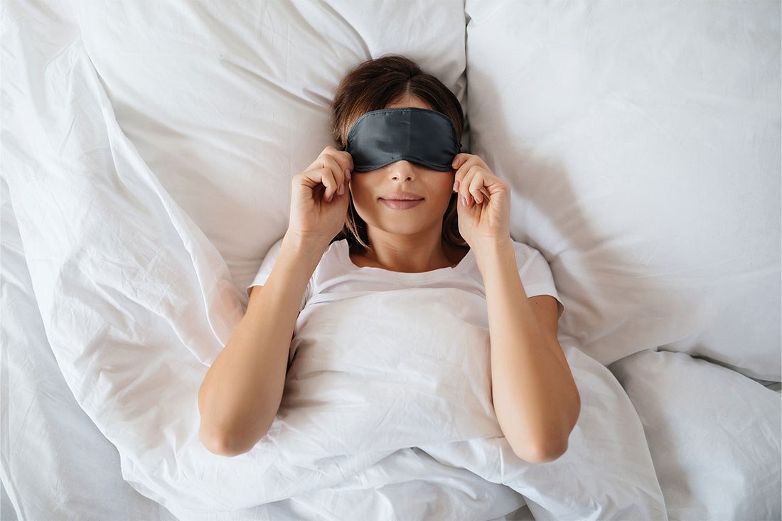 woman laying in bed with white sheets with black sleeping mask over eyes