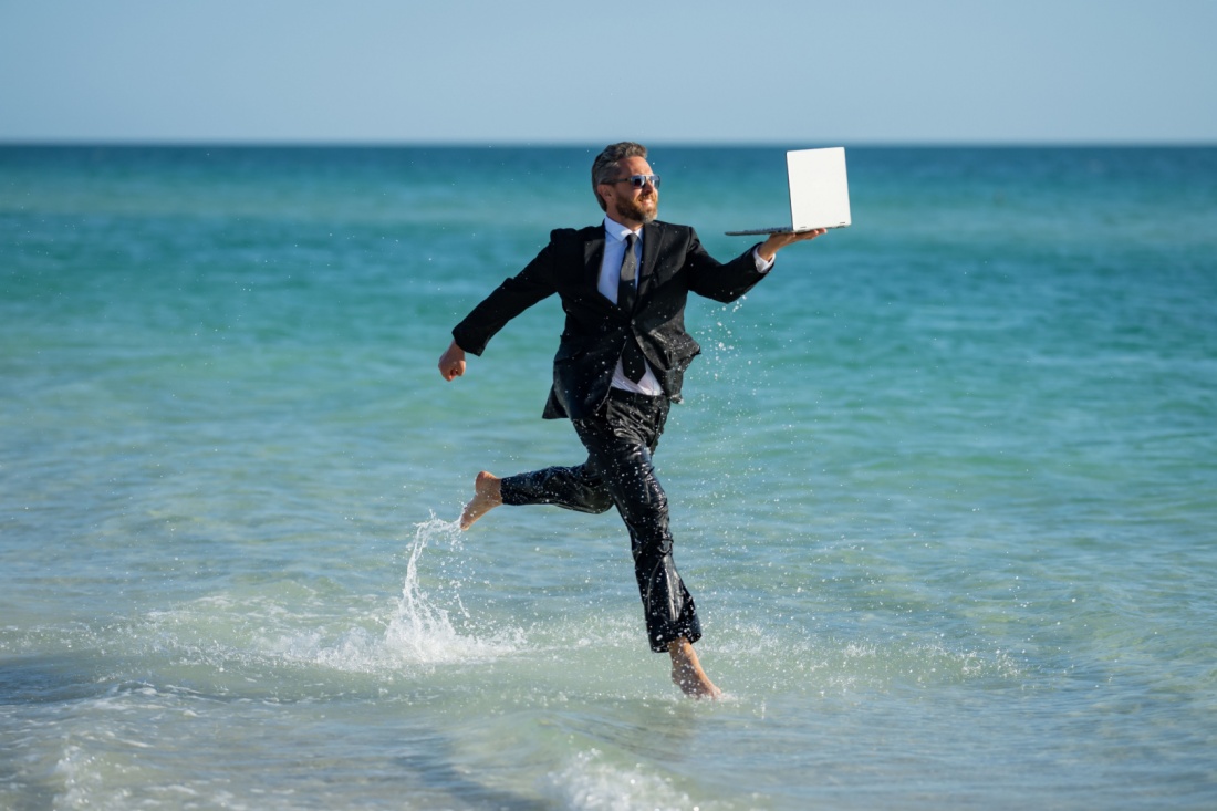 A businessman in a full suit and tie frolics across the beach with his laptop like a LinkedIn lunatic