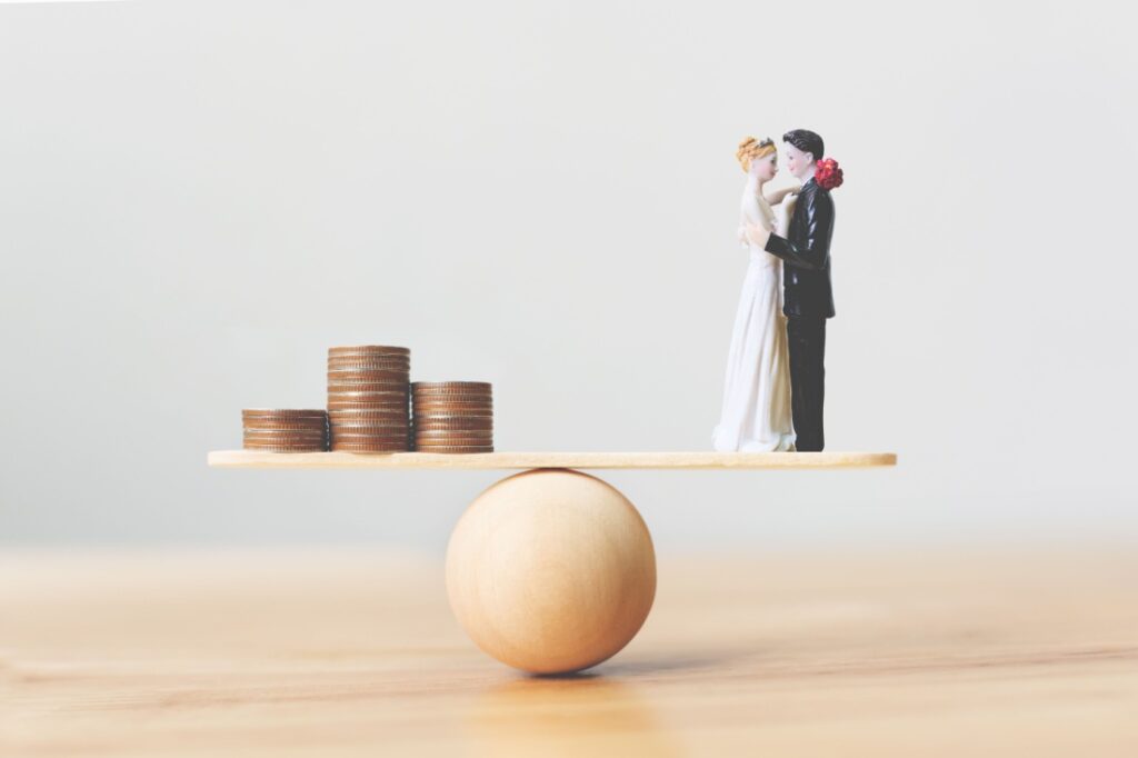 A figure of a bride and groom balanced on a scale against a stack of coins.