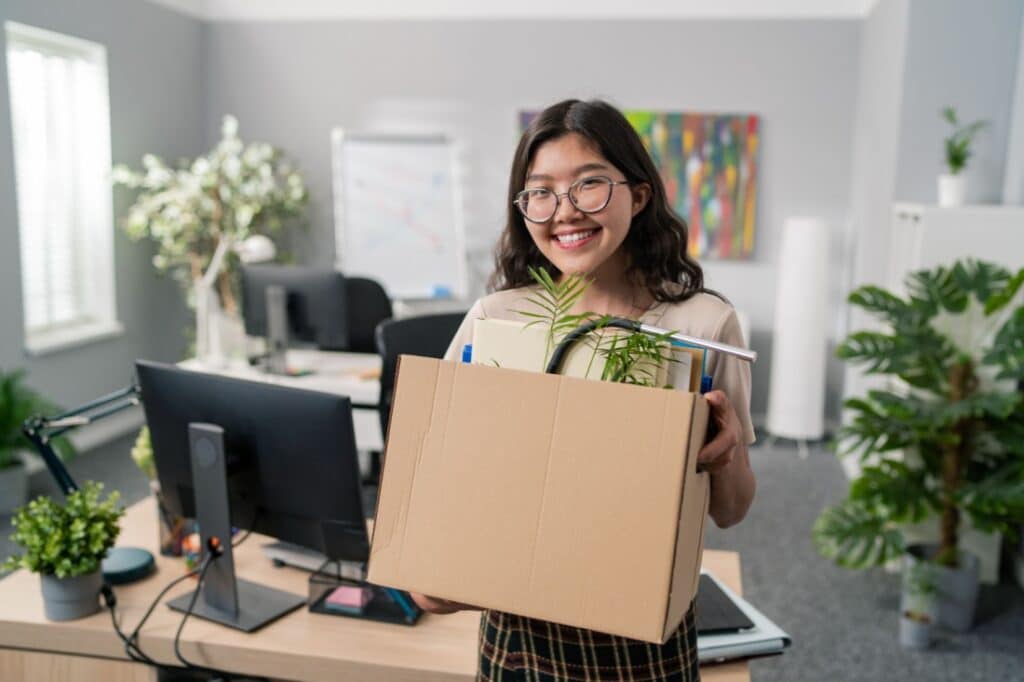 A smiling woman carries a box of her desk supplies out of an office