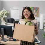 A smiling woman carries a box of her desk supplies out of an office