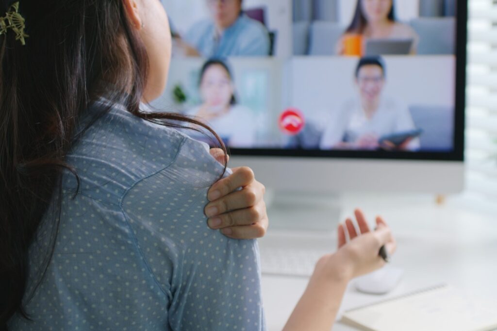 An anxious woman grabs her shoulder while facing a computer monitor showing a zoom meeting.