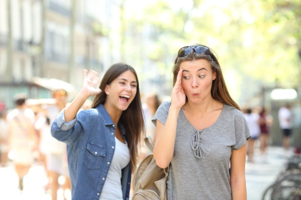 A woman avoids making eye contact with another woman who is waving enthusiastically at her