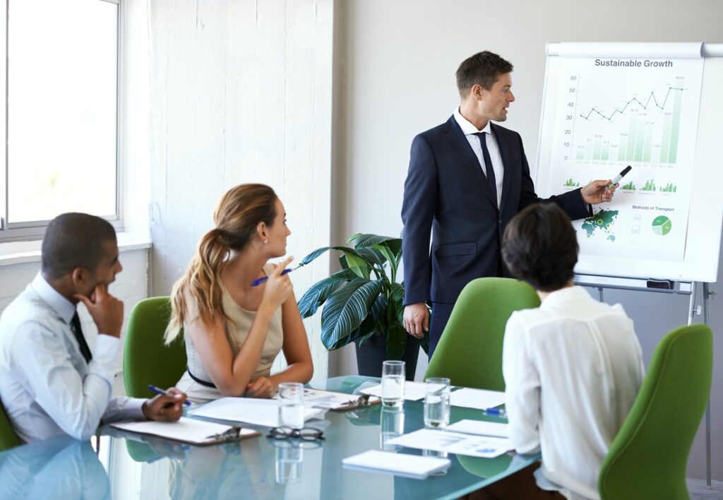 people sitting in a meeting with one person standing at a presentation board