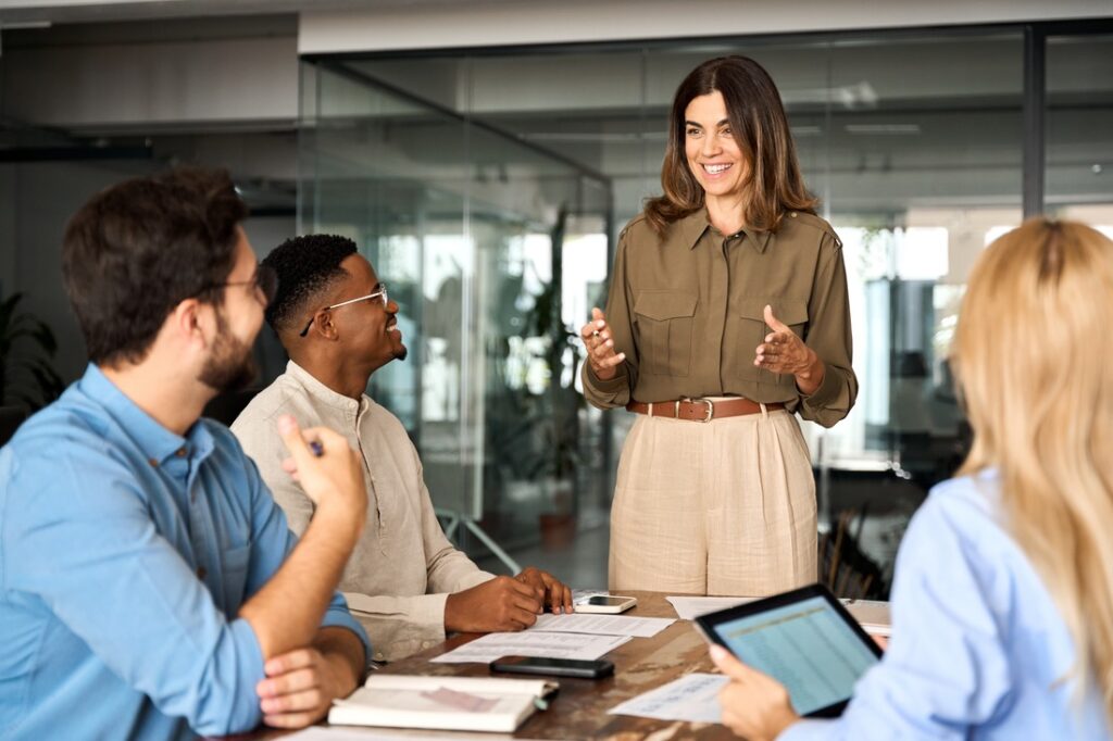 a boss speaking to employees who are sitting at a table at their place of work