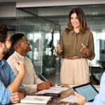 a boss speaking to employees who are sitting at a table at their place of work