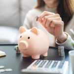 Young woman saving money monthly expenses putting coin in to piggy bank on the table.