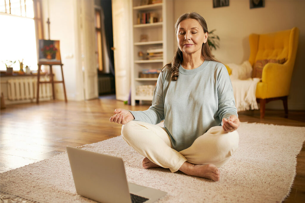 senior woman sitting on carpet in front of open laptop keeping eyes closed and legs crossed