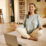 senior woman sitting on carpet in front of open laptop keeping eyes closed and legs crossed