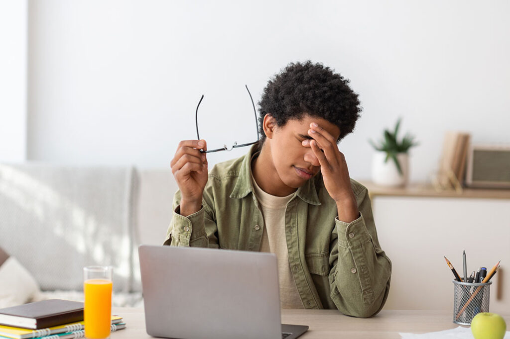 Black teenager feeling screen fatigue, rubbing irritated eyes, sitting at desk with laptop