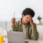 Black teenager feeling screen fatigue, rubbing irritated eyes, sitting at desk with laptop