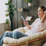 Young woman wears casual clothes sits in armchair with headphones