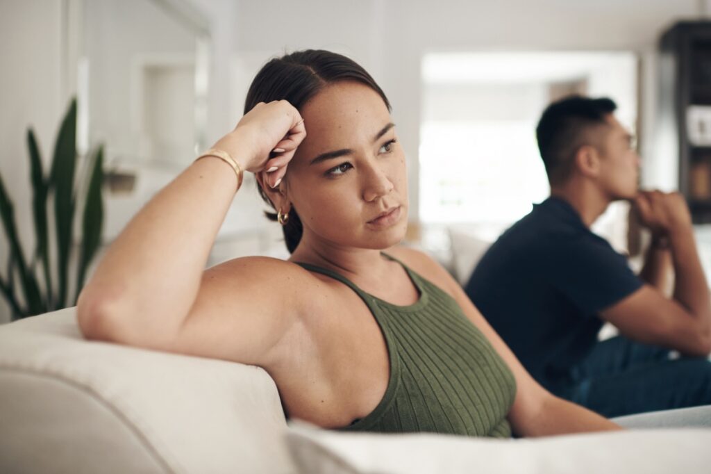 A woman sits on a couch, looking frustrated, turned away from her partner.