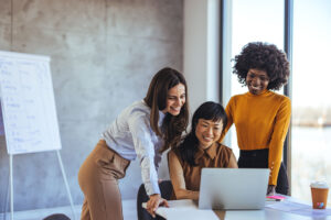 Three women of different ethnicities smiling and working together at a laptop in a modern office.