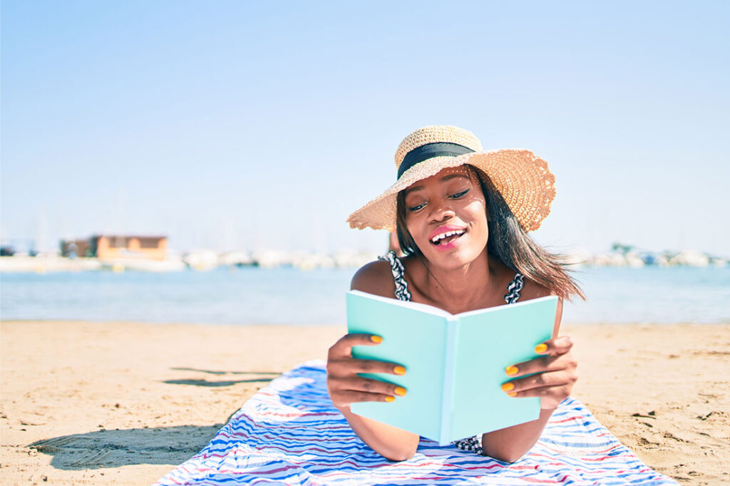 Young african american woman on vacation laying on the towel reading book at the beach