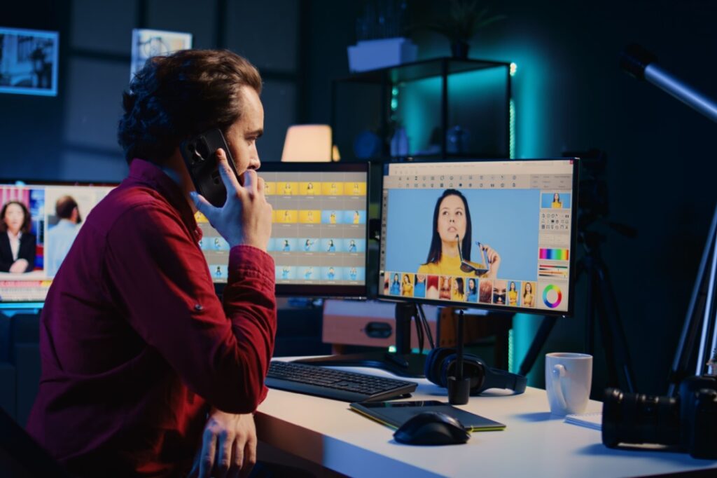 A photo editor talks on the phone while sititng in front of a multi-monitor editing set-up.