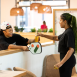 A woman gives a beverage to another woman holding a pickleball racquet.