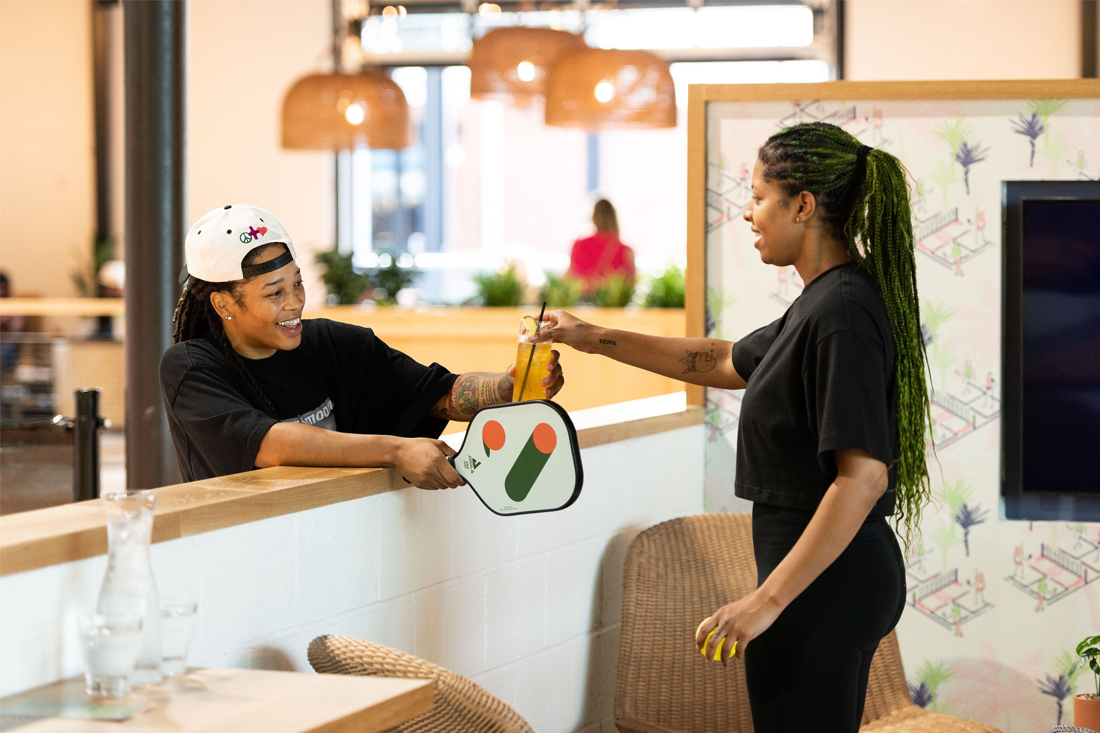 A woman gives a beverage to another woman holding a pickleball racquet.