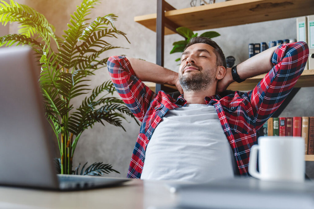 Middle aged man resting in front of laptop screen at his work place, with arms behind the head focused