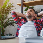 Middle aged man resting in front of laptop screen at his work place, with arms behind the head focused