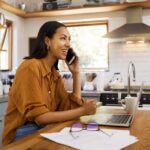 woman sitting at a table in her kitchen with her laptop on the table in front of her while she is talking on the phone