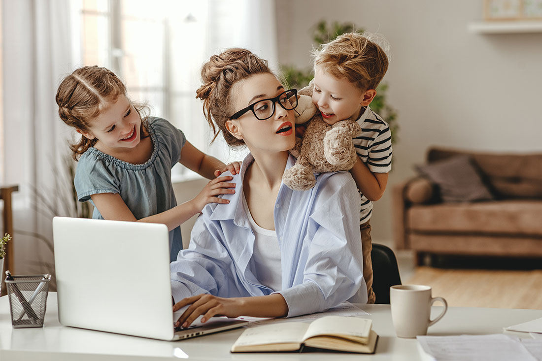 girl hugging and small son showing toy to busy mother sitting at table with cup of coffee and using laptop against blurred interior of modern apartment