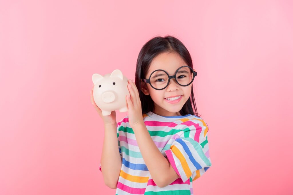 A little girl with large glasses smiles while holding up a piggy bank against a pink background.