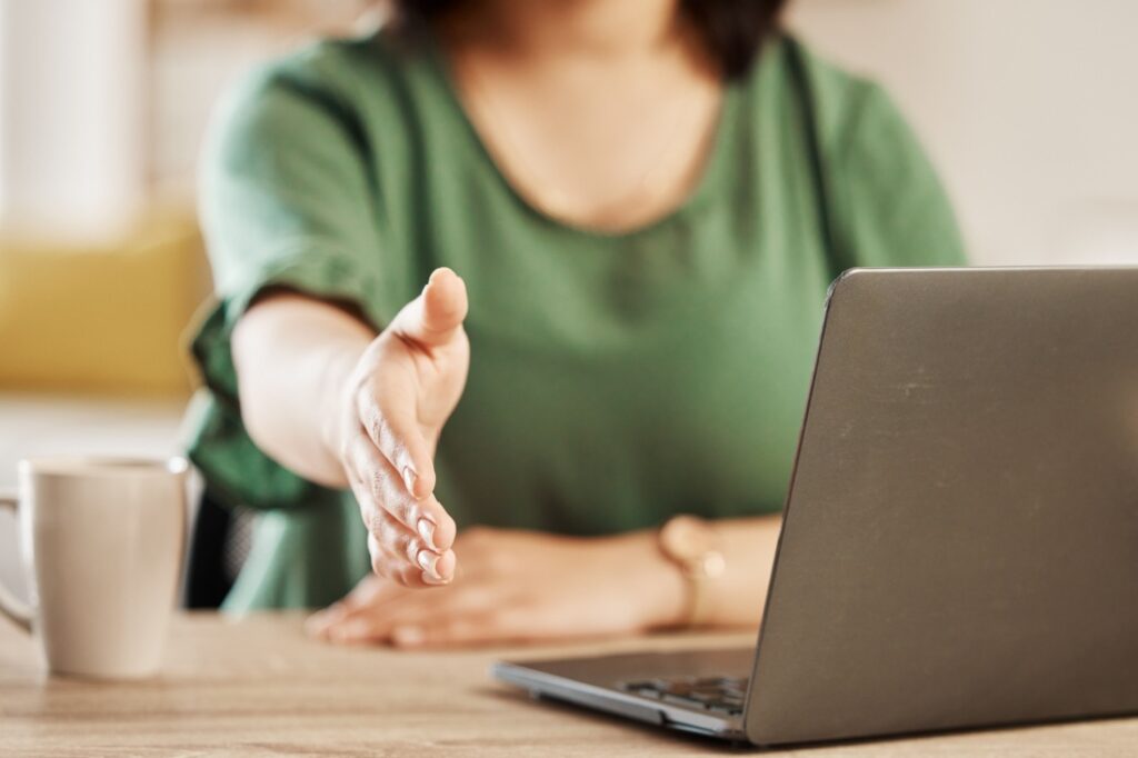 A woman reaches out her hand for a handshake while sitting in front of a laptop.