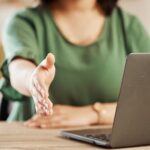 A woman reaches out her hand for a handshake while sitting in front of a laptop.