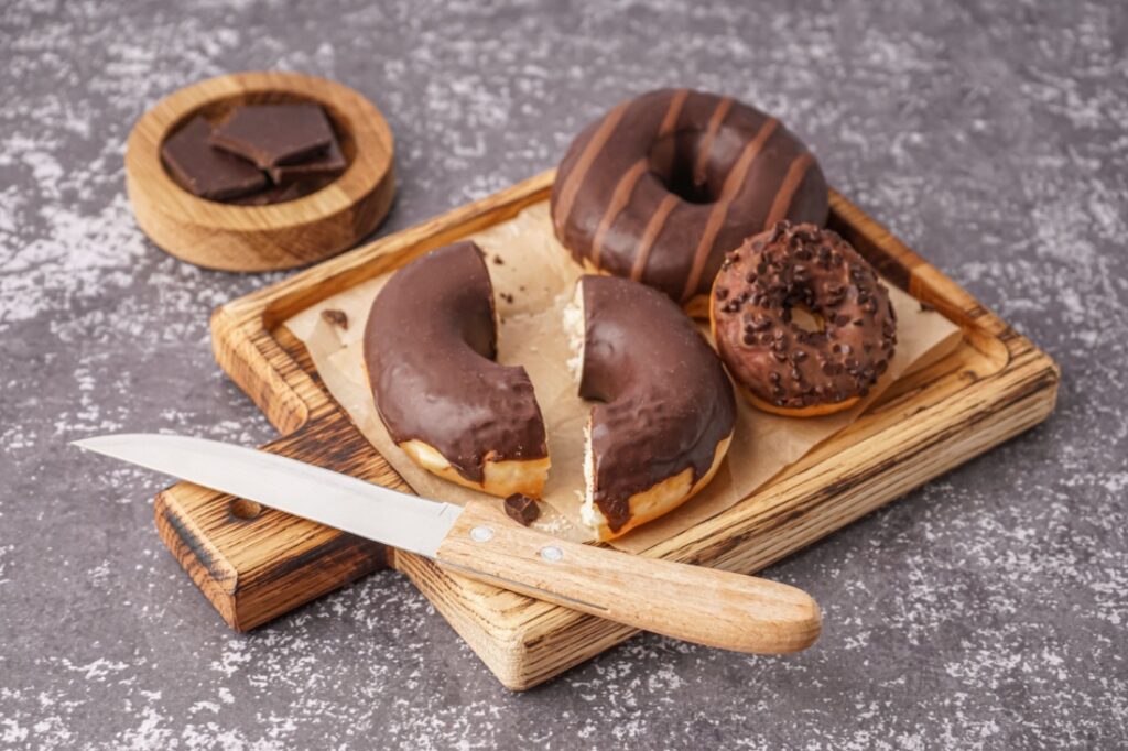 A steak knife on a chopping block next to three chocolate donuts