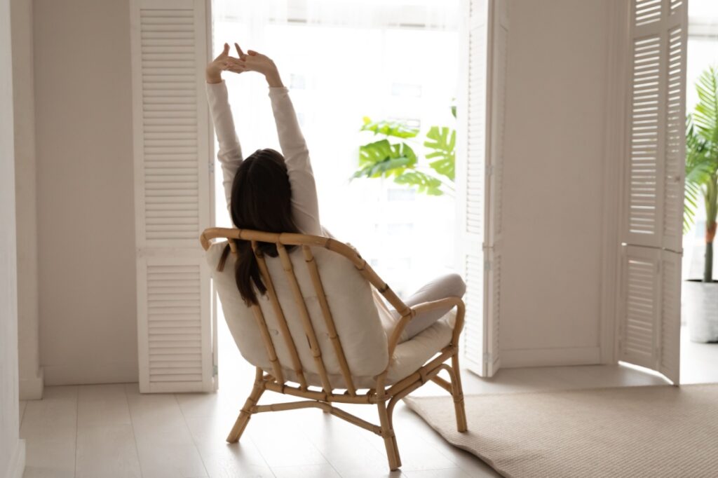 A woman stretches while sitting in a chair facing a window.